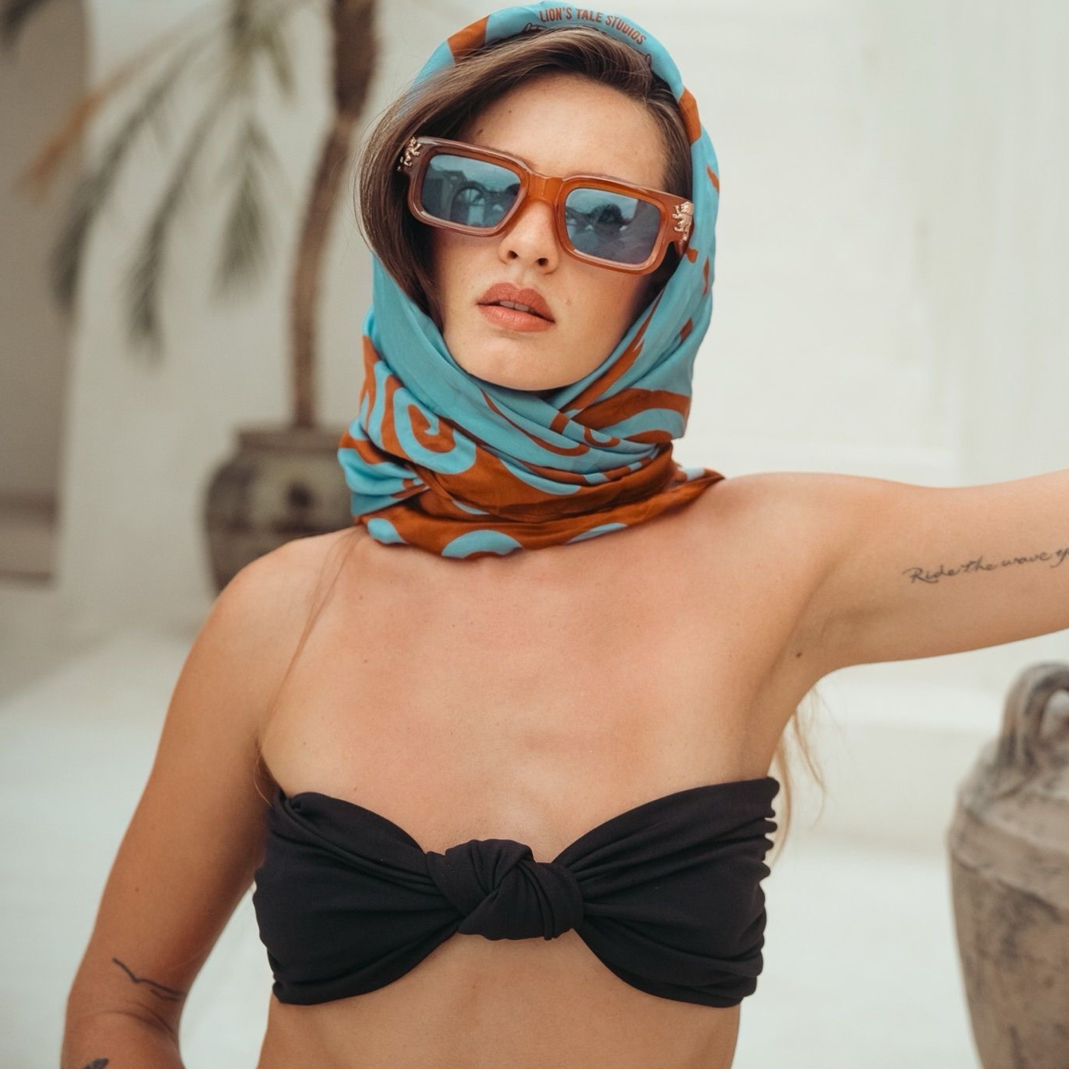 Tattooed female model wearing a 100% pure silk scarf by Lionâs Tale Studios â luxury babushka wrap with Moluccan ancestral symbols, cream and oxford blue design, hypoallergenic and breathable silk, styled for unisex minimalist fashion and heritage streetwear. Vrouwelijk model met tattoos draagt 100% zijden sjaal van Lionâs Tale Studios â luxe babushka-stijl met Molukse symbolen, crĂšme en oxfordblauw, hypoallergeen, geschikt voor uniseks mode en erfgoed-geĂŻnspireerde. masami bouwman leimena eyewear babushka